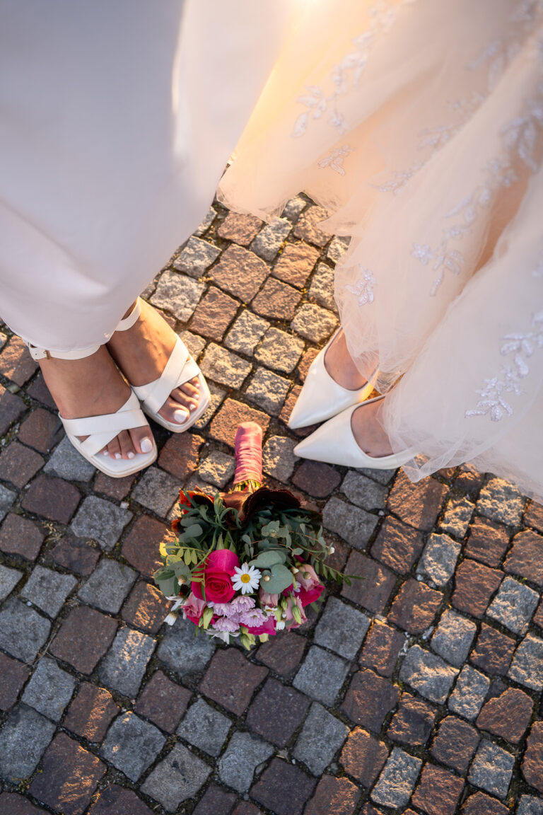 Brautstrauß mit margariten und pinken Blumen liegt auf dem Boden neben den Füßen zweier Bräute