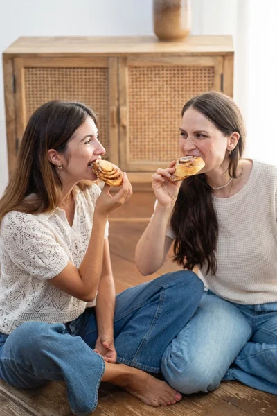 Zwei Frauen sitzen auf Boden vor Schrank und essen Zimtschnecken