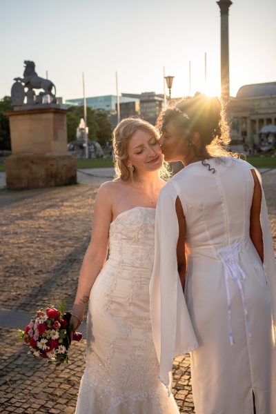Braut küsst blonde Frau im Hochzeitskleid auf die Wange zur goldenen Stunde auf dem Stuttgarter Schlossplatz